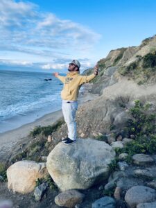 Photo of student Zachary Olivan standing on a rock at the coast with arms outstretched looking up torwards sky and facing ocean with a peaceful look on his face.